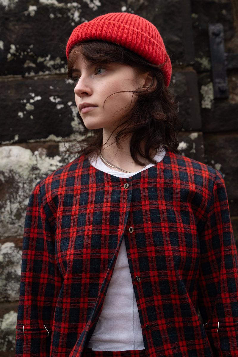 Portrait of a woman wearing a red beanie and red plaid jacket against an old stone wall background.