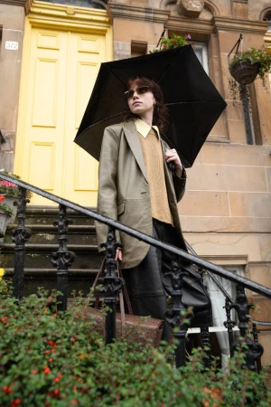 Model standing on stone steps holding a black umbrella wearing a blazer and sweater with a yellow door in the background
