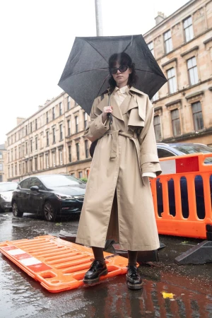 Woman in beige trench coat holding a dark umbrella standing on a wet city street with parked cars and orange barriers.