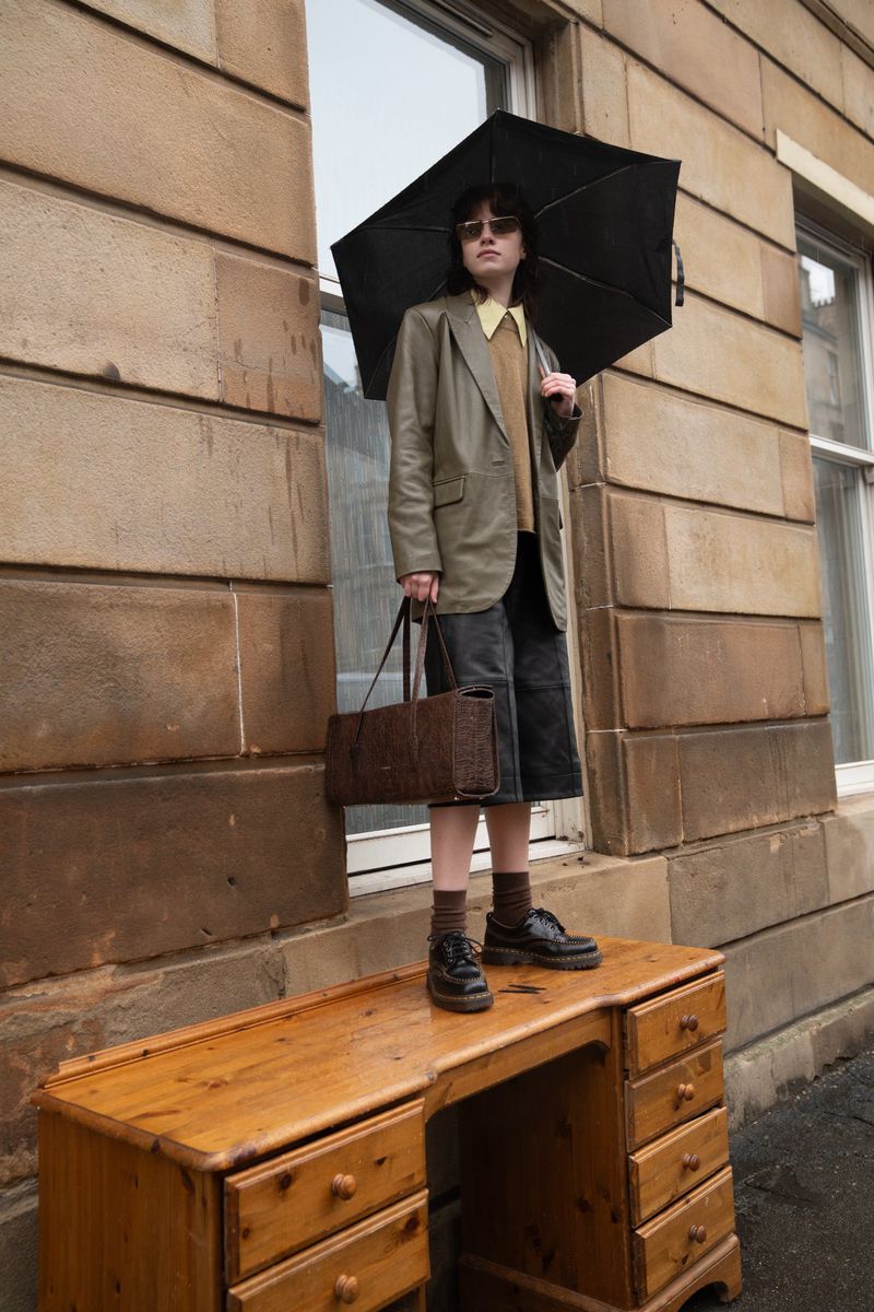 Model standing on a wooden desk holding an open black umbrella in front of a stone building exterior for Flanelle Magazine