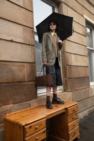 Model standing on a wooden desk holding an open black umbrella in front of a stone building exterior for Flanelle Magazine