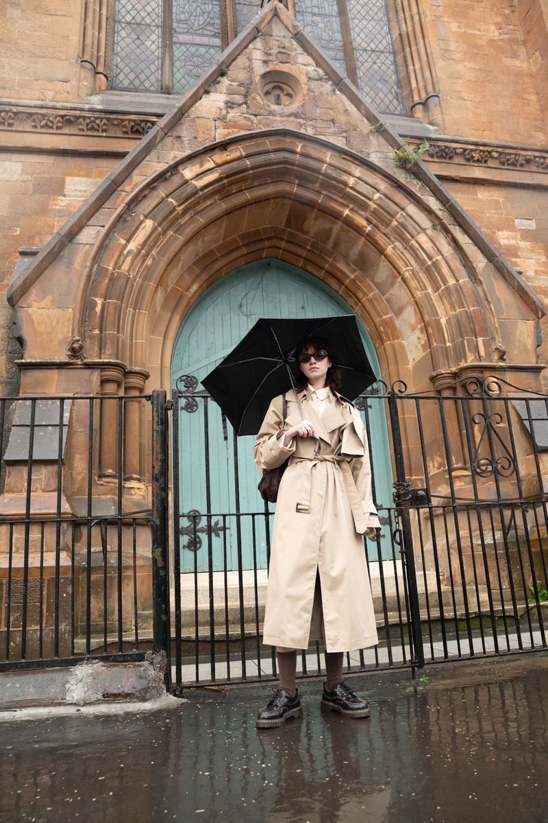 Model wearing beige trench coat holding black umbrella in front of a stone building exterior with blue door