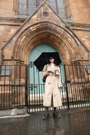 Model wearing beige trench coat holding black umbrella in front of a stone building exterior with blue door