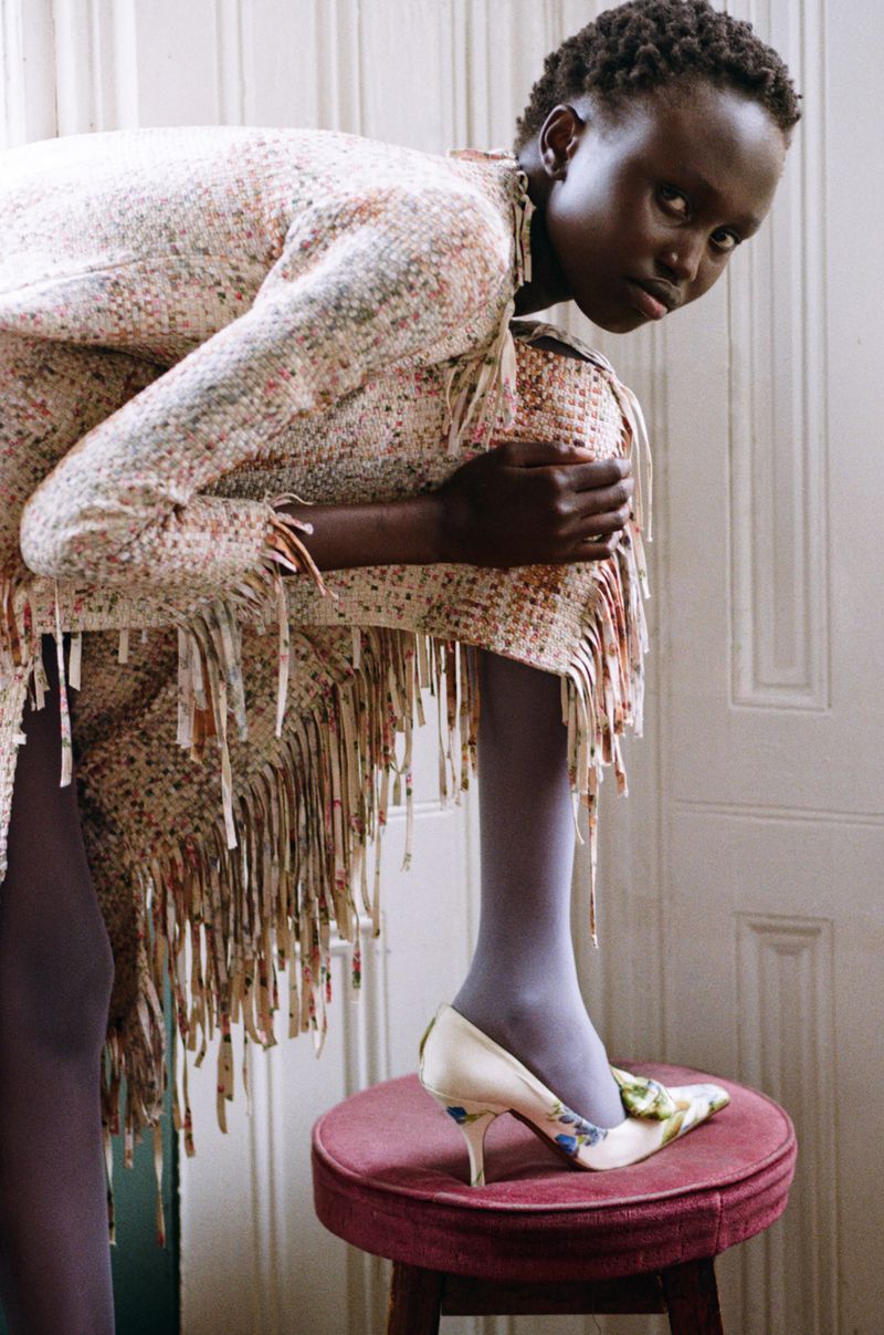 Model in a fringed tweed dress and heels posing over a small red velvet stool against a paneled wall.