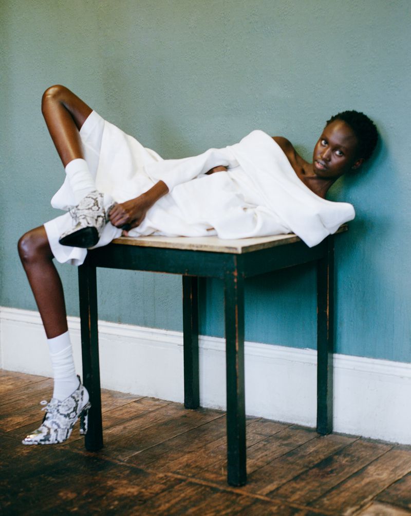 Woman in white outfit with patterned heels poses on table against muted wall, capturing a modern fashion editorial look.