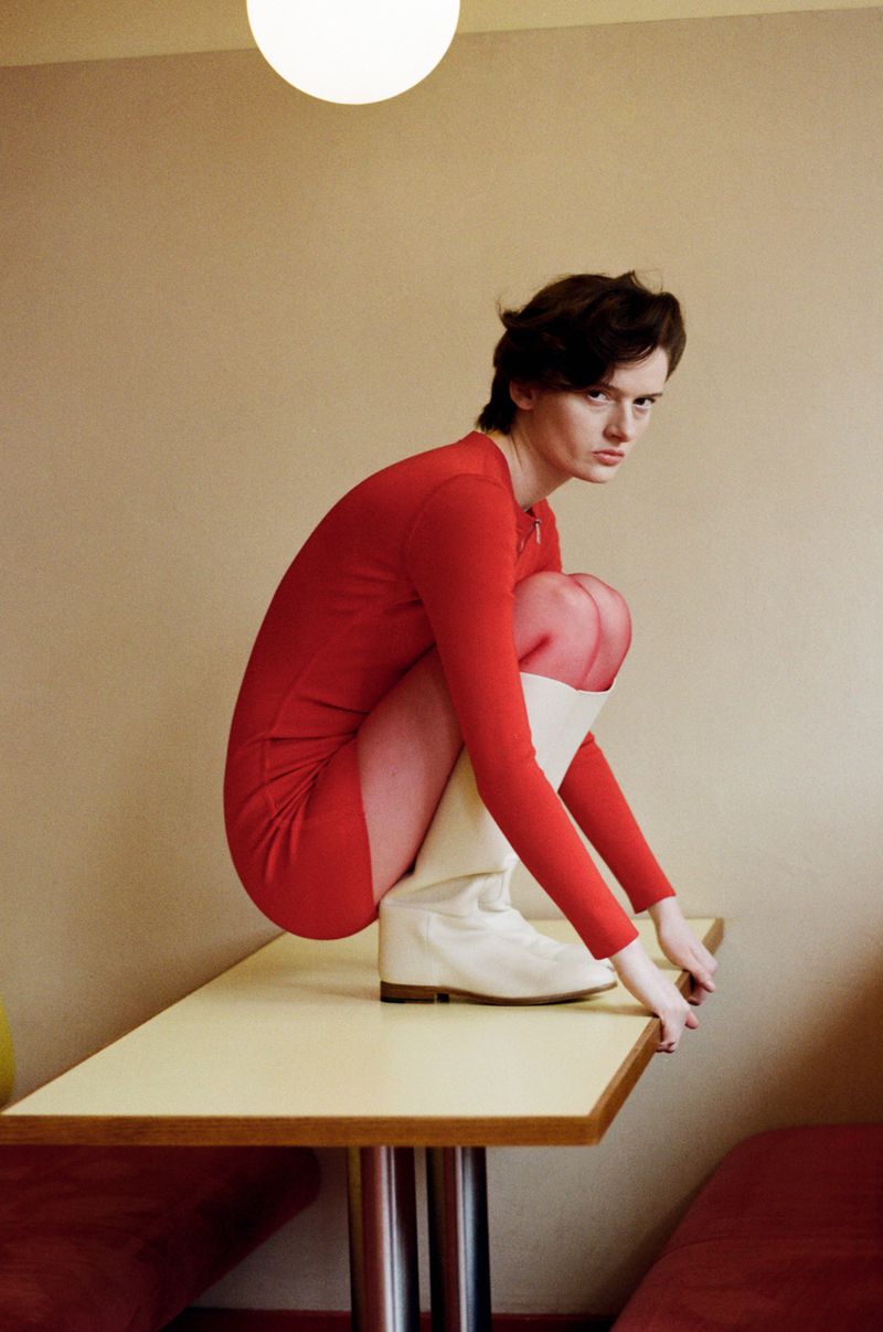 Model wearing a red dress and white boots crouching on a table inside a diner
