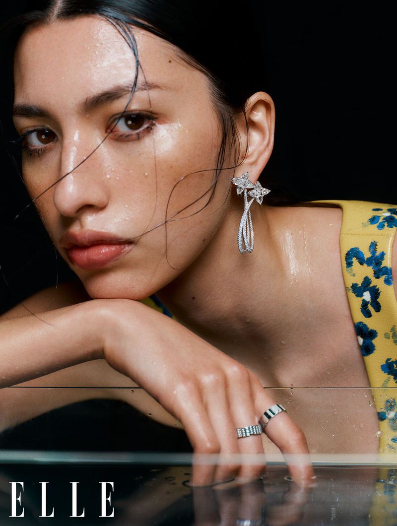 Close-up portrait of a woman resting her chin on her hand, wearing floral yellow dress and diamond earrings against a dark background.