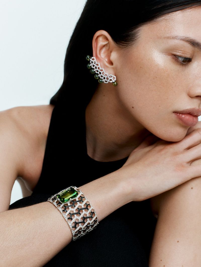 Close-up portrait of a model wearing an intricate diamond and green gemstone ear cuff and cuff bracelet against a plain background