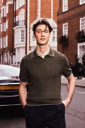 A stylish man poses on a London street wearing a green collared shirt, navy pants, and a patterned neck scarf, with brick buildings in the background.