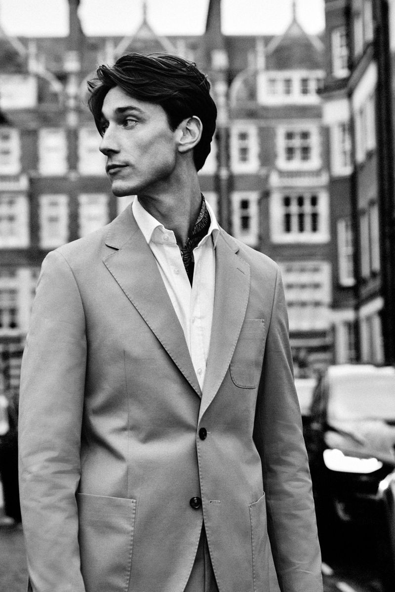 Monochrome portrait of a male model in a tailored suit and open collar shirt outdoors on a city street.