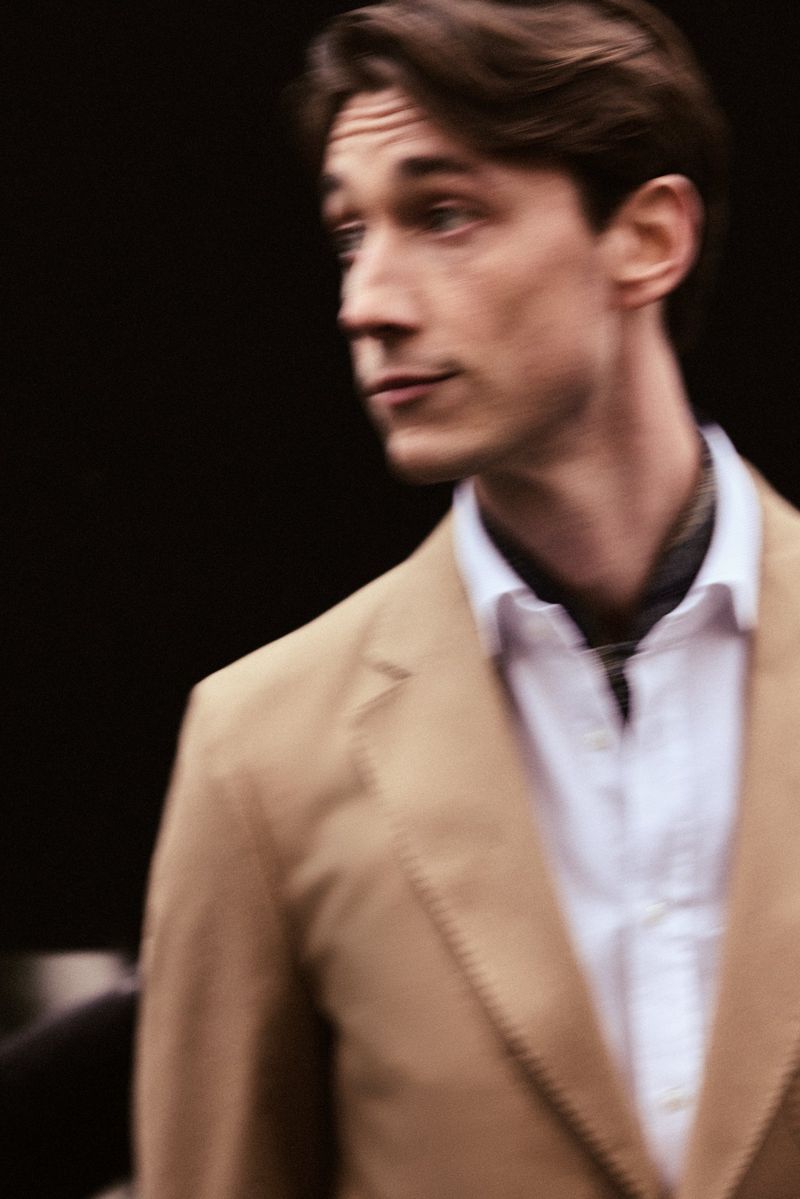 Man wearing a camel blazer, white shirt, and dark scarf against a dark background in the Carolina Herrera Matryoshka Pocket campaign.