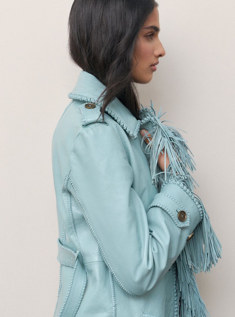 Profile view of a woman wearing a light blue fringed leather jacket against a seamless off-white background.