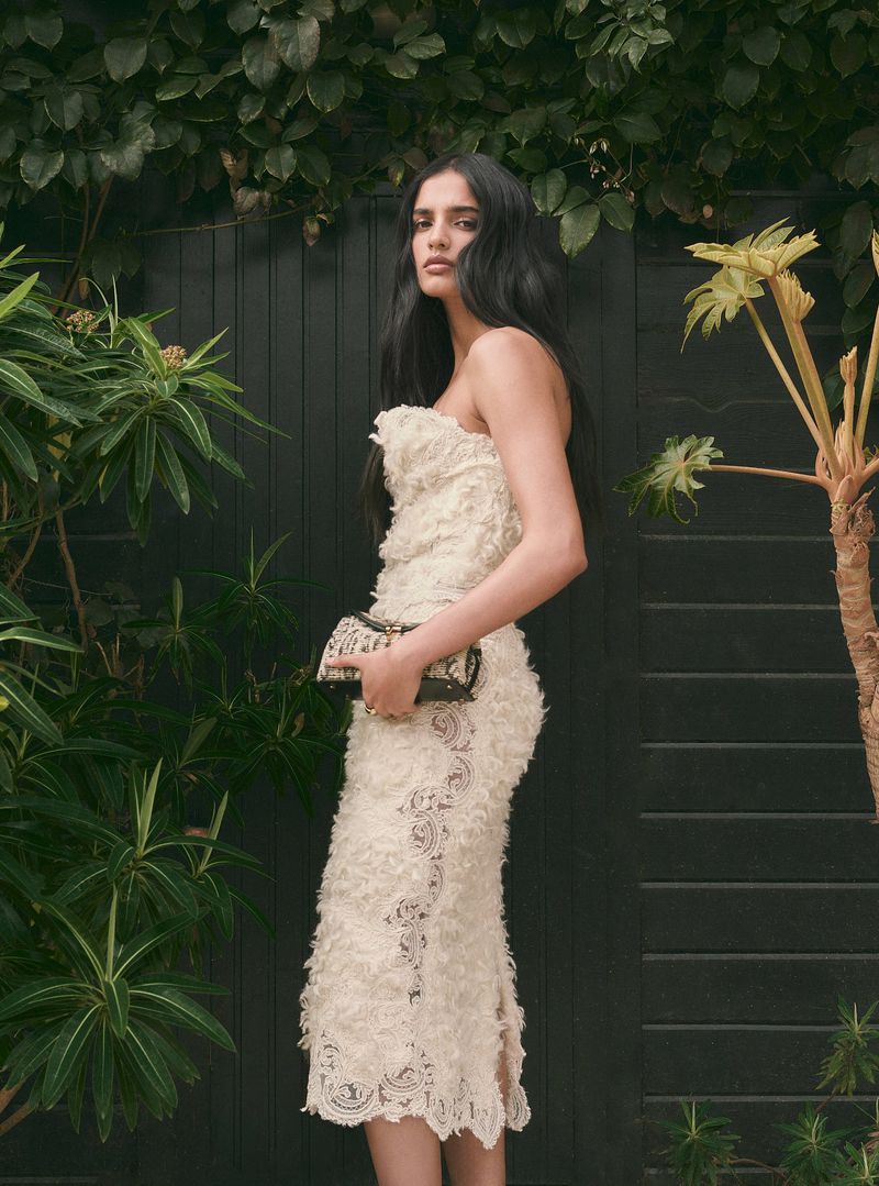Model in a cream strapless textured dress holding a small clutch against a dark wooden fence and foliage.