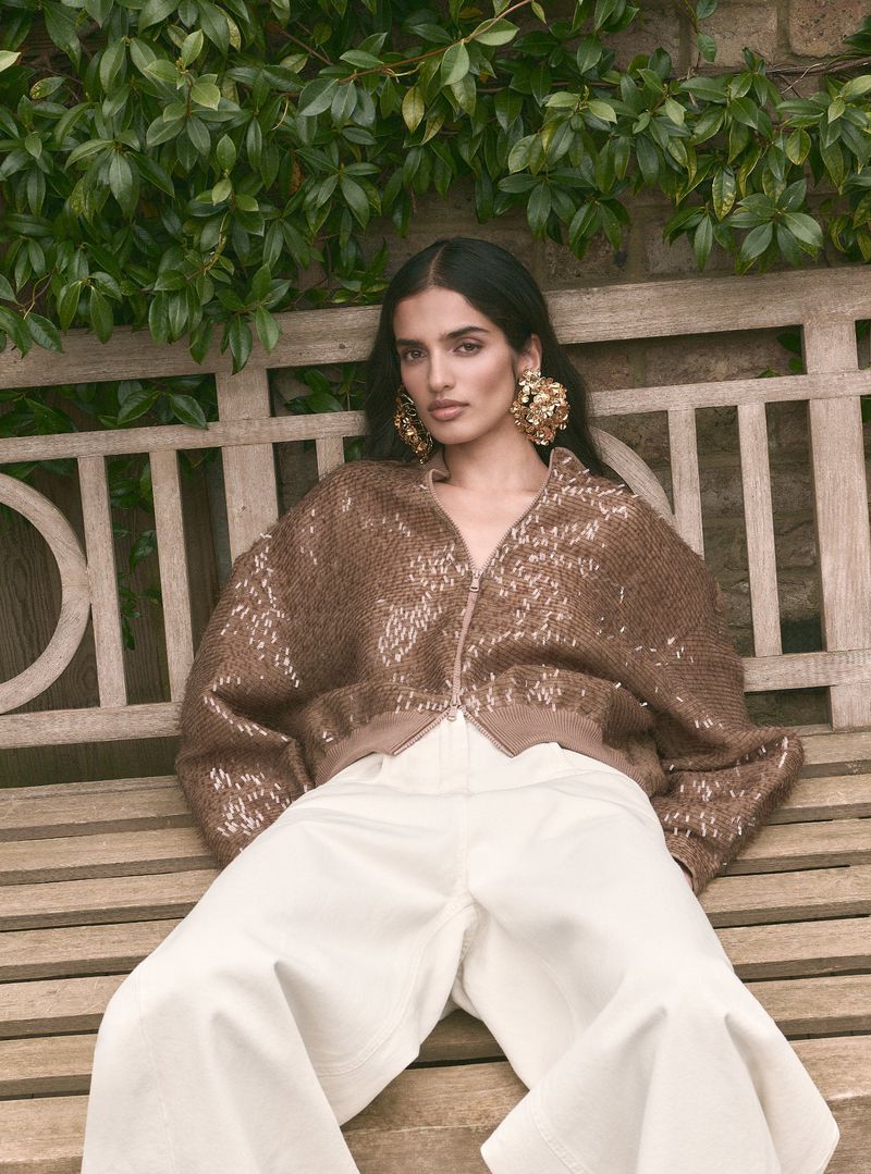 Model wearing a textured brown jacket and white trousers seated on a wooden garden bench with foliage background.