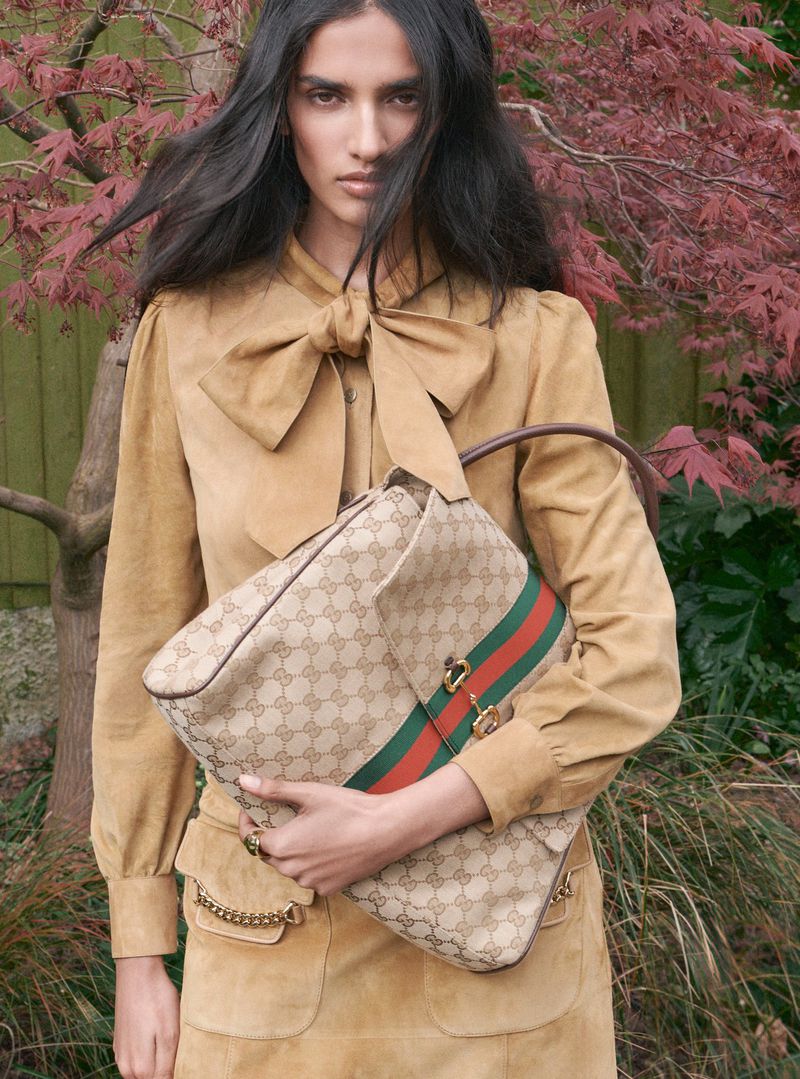 Model wearing a tan suede bow blouse holding a monogram handbag outdoors before reddish foliage