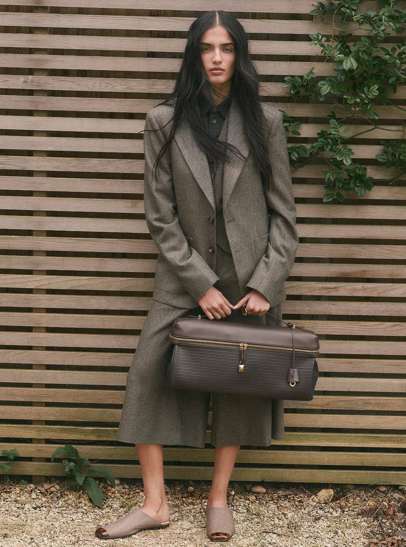 Model Anjali Torvi in a grey tweed suit holding a dark leather bag against a horizontal wooden slat fence