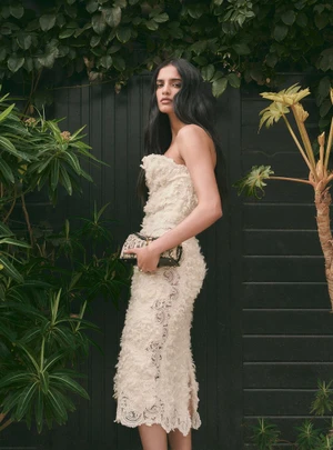 Model in a cream strapless textured dress holding a small clutch against a dark wooden fence and foliage.