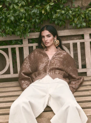 Model wearing a textured brown jacket and white trousers seated on a wooden garden bench with foliage background.