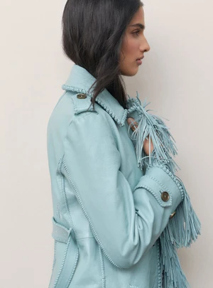 Profile view of a woman wearing a light blue fringed leather jacket against a seamless off-white background.