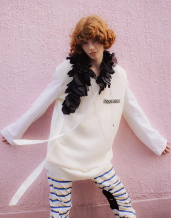 A red-haired person poses in a cream-colored outfit with a feathered boa against a textured pink wall, showcasing unique style and fashion.
