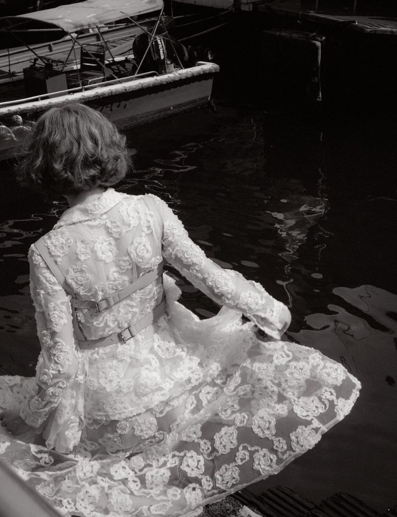 Back view of a woman in a floral lace dress sitting by the water with boats in the background, in a monochrome photograph.