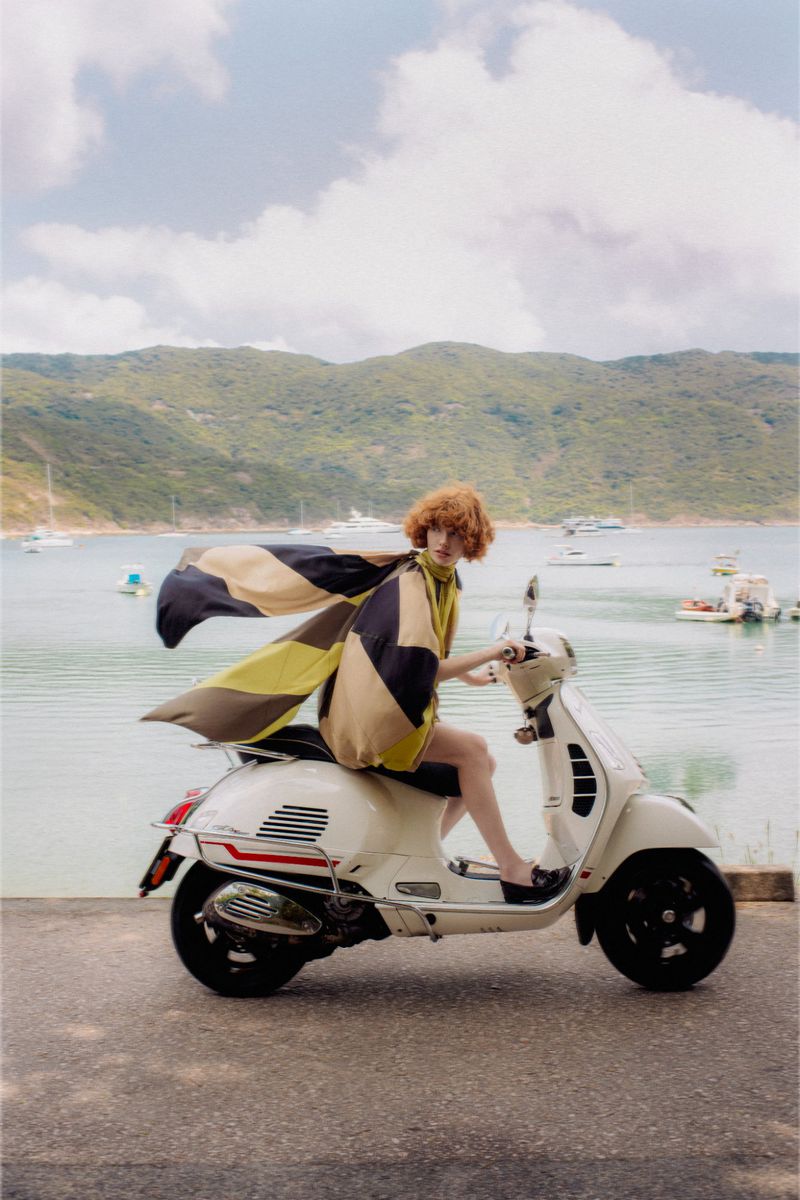 A woman with red hair and a patterned cape rides a white vintage scooter on a road with the sea and green hills in the background.