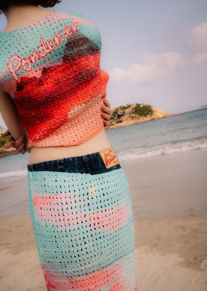 Young woman on a beach wearing a colorful, crocheted outfit, standing near the ocean on a sunny day with a small island in the background.