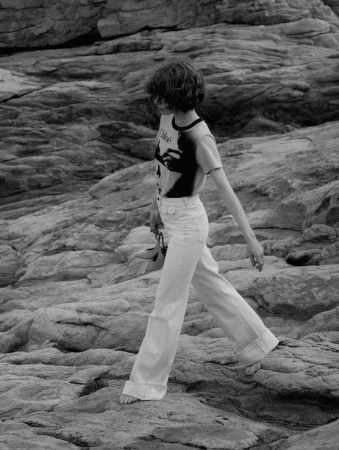 Monochrome fashion shot of a young woman in white pants walking barefoot on rocks, holding shoes, wearing graphic tee.