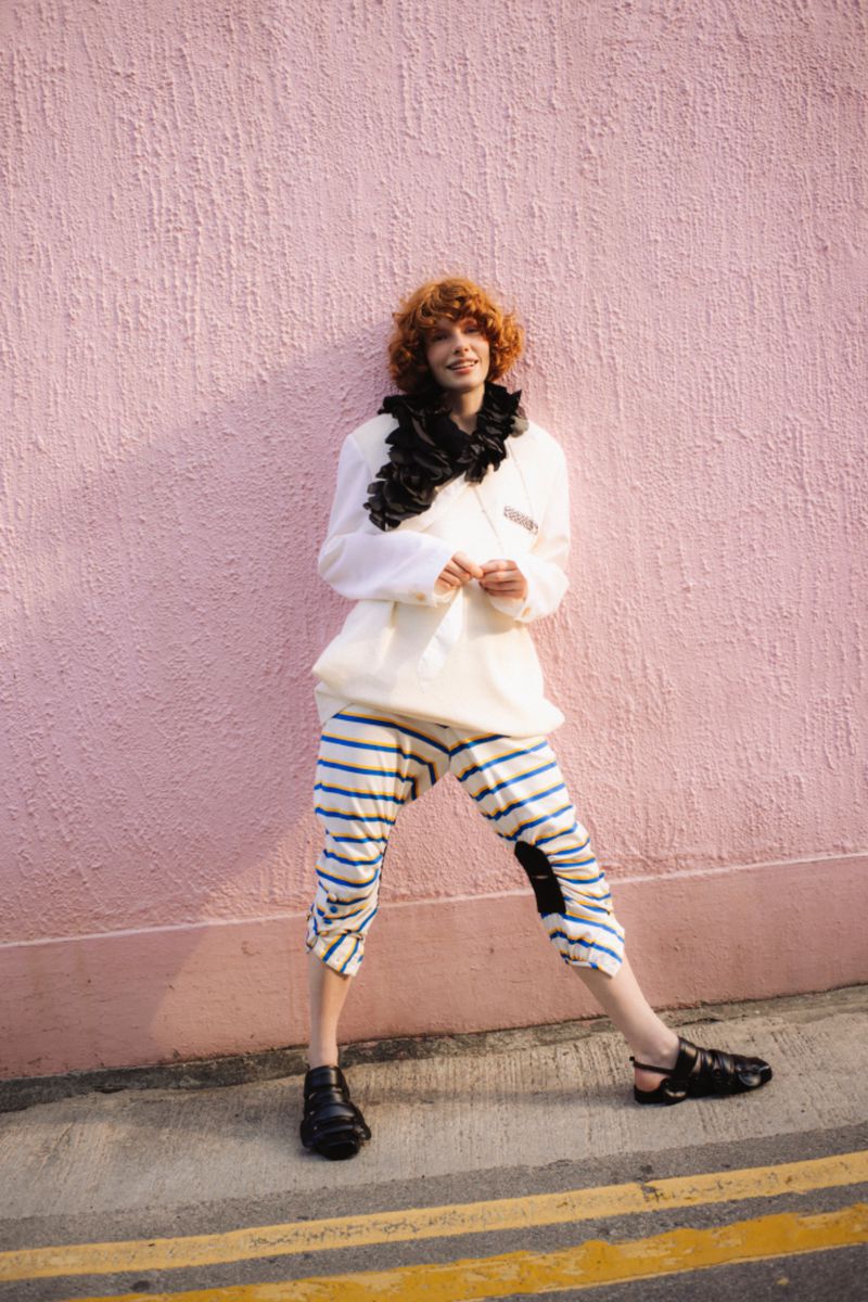 A person with curly red hair stands confidently against a textured pink wall wearing oversized white top, striped pants, and black shoes.