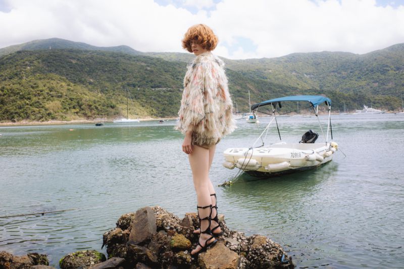 Woman in a shaggy top stands on rocks near the water's edge, with a small boat floating in the background on an overcast day.