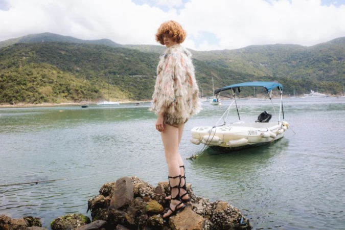 Woman in a shaggy top stands on rocks near the water's edge, with a small boat floating in the background on an overcast day.