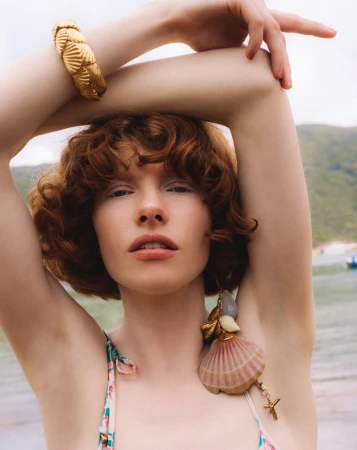 Close-up portrait of a young woman with red curly hair wearing gold jewelry, standing on a sandy beach with a hillside view in the background.