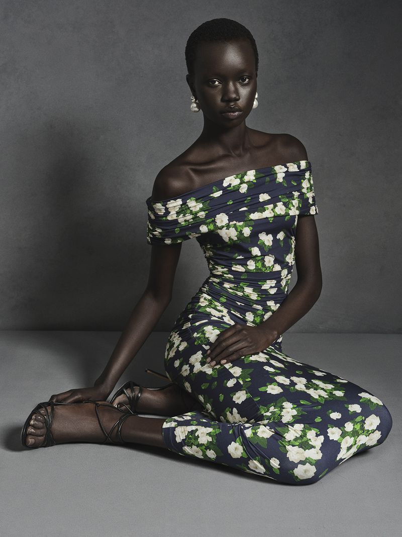 Black model seated on the floor wearing an off-the-shoulder floral print dress against a dark studio background