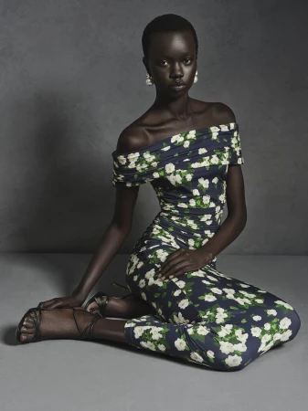 Black model seated on the floor wearing an off-the-shoulder floral print dress against a dark studio background