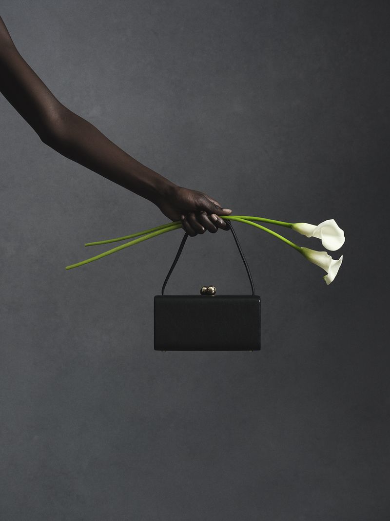 A dark-skinned person holding a black handbag and white calla lilies against a textured grey background.
