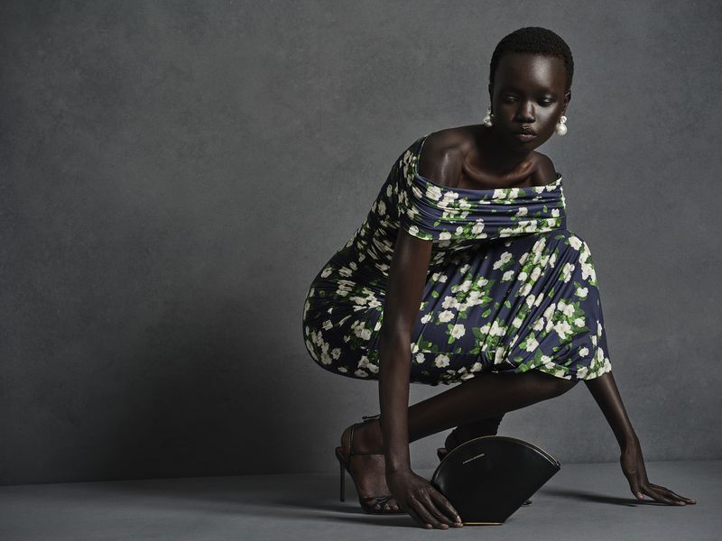 Black woman crouching in an off-the-shoulder floral print dress with a black handbag against a grey textured backdrop