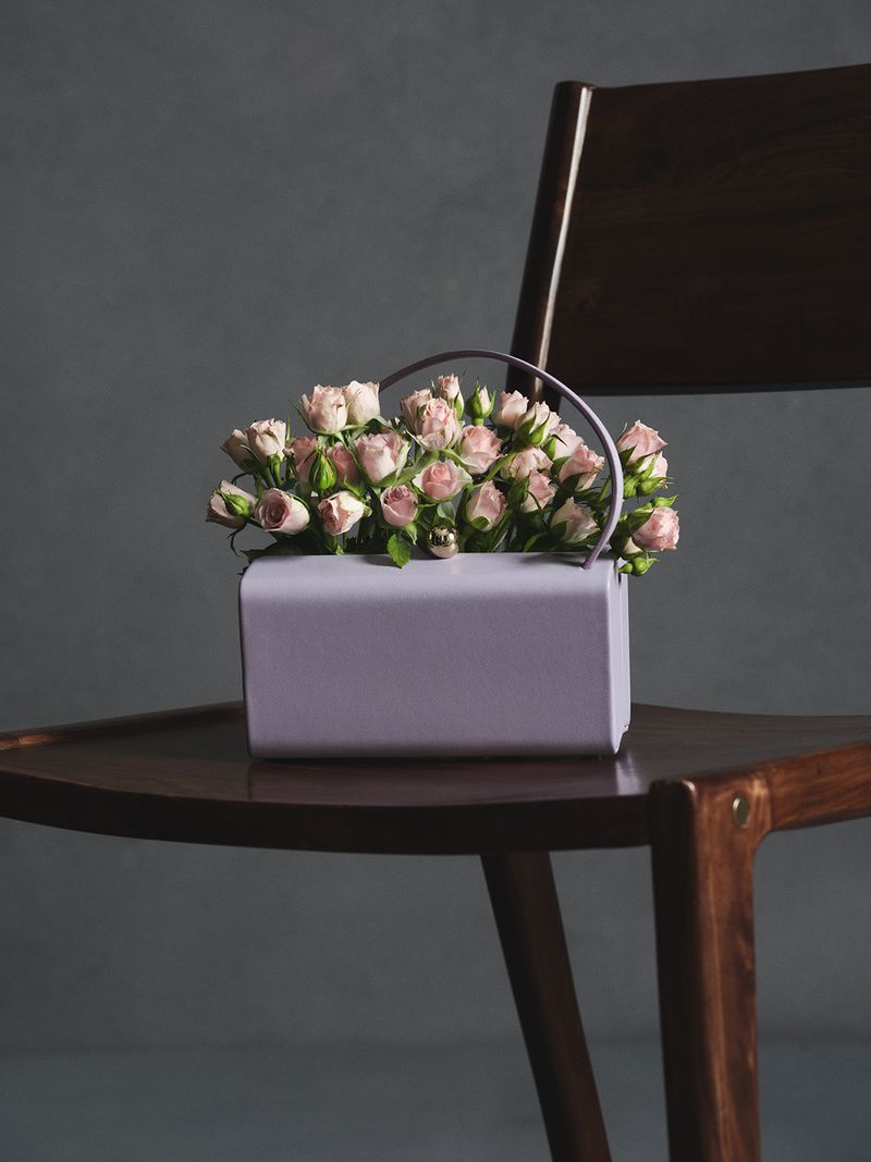 A lilac handbag filled with pale pink miniature roses resting on a dark wooden chair