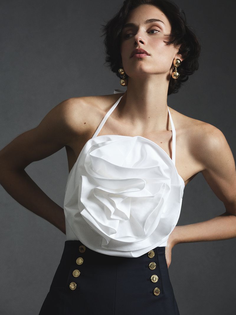 Model wearing a white halter top with a large floral ruffle and dark bottoms against a grey background.
