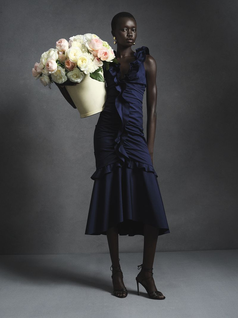 Model in a navy blue ruffled dress holding a large pale bucket of roses against a grey studio backdrop.