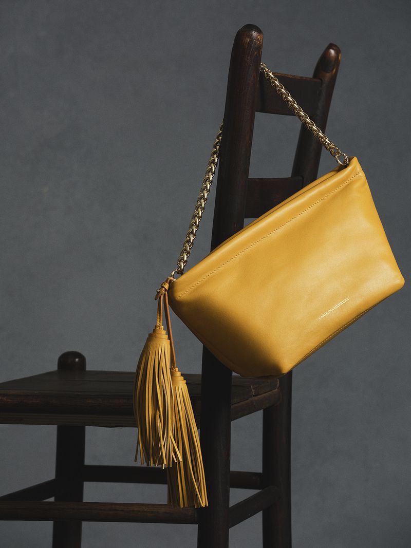 Bright yellow leather shoulder bag with a gold chain strap hanging on a dark wooden chair against a grey textured studio backdrop.
