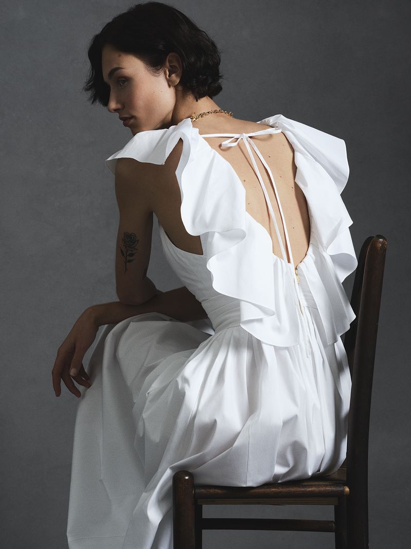 Model seated backwards on a wooden chair wearing a white ruffled dress for Carolina Herrera Summer 2026
