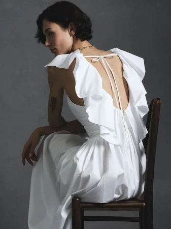 Model seated backwards on a wooden chair wearing a white ruffled dress for Carolina Herrera Summer 2026