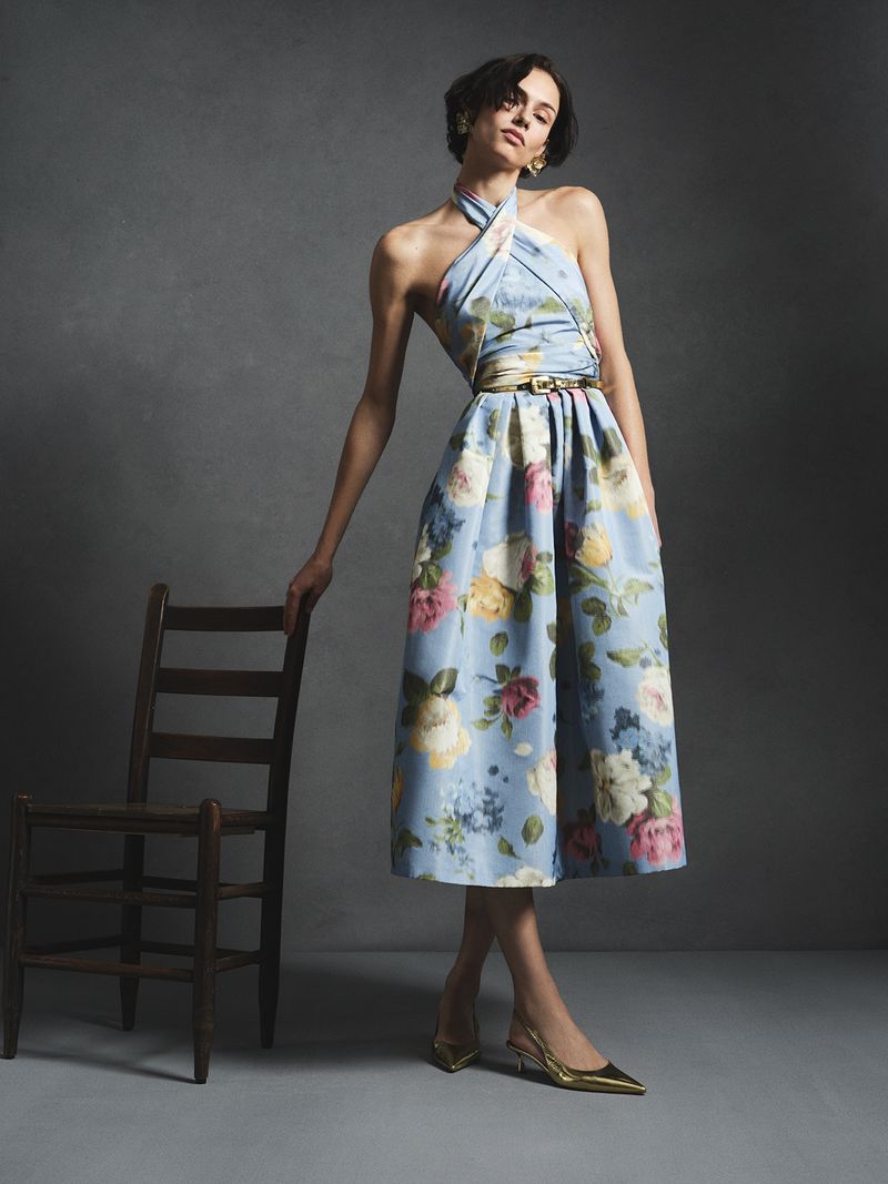 Model in a light blue floral halter dress resting hand on a dark wooden chair against a muted grey studio backdrop.