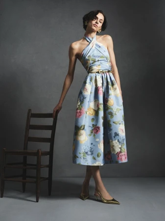 Model in a light blue floral halter dress resting hand on a dark wooden chair against a muted grey studio backdrop.