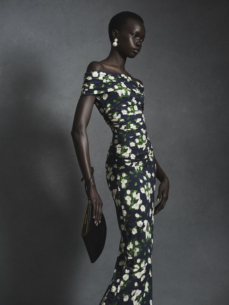 Model in off-the-shoulder floral dress holding a black clutch against a textured grey studio background