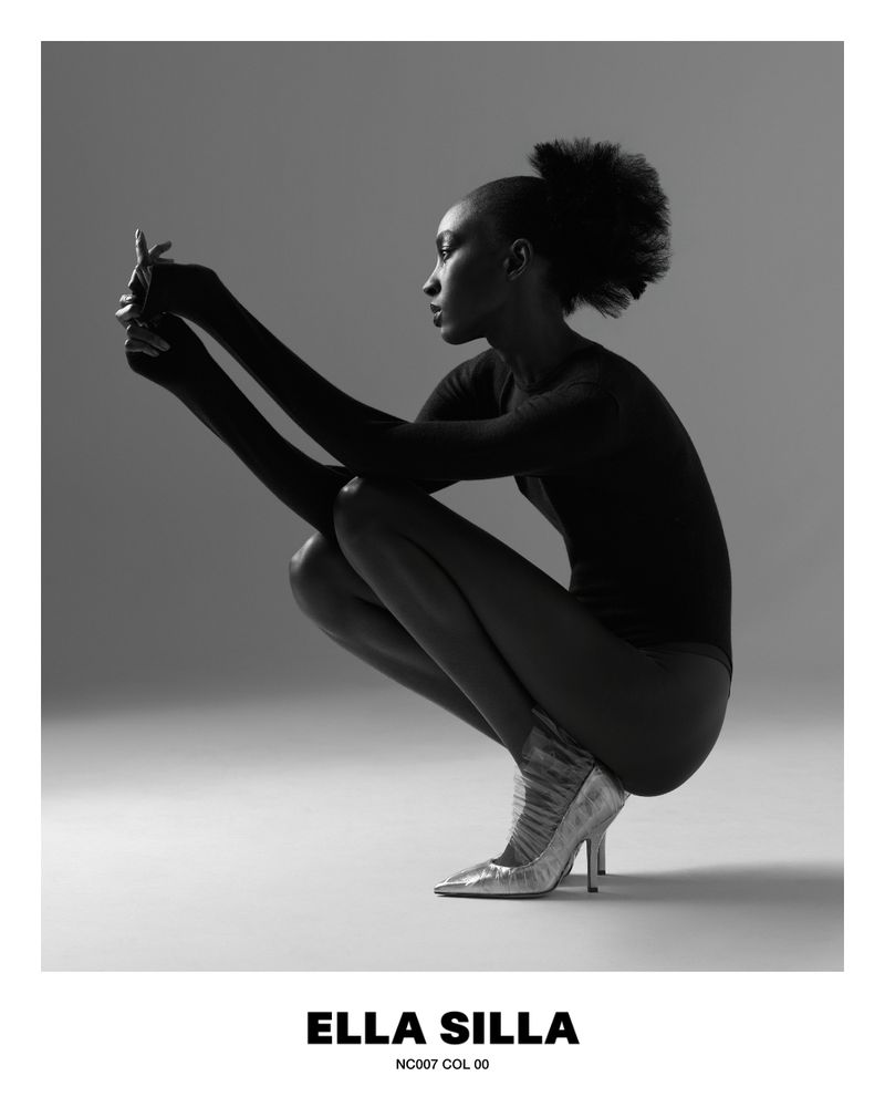 Black and white studio shot of an African-American woman in a bodysuit and high heels, crouching and looking at something in her hands.