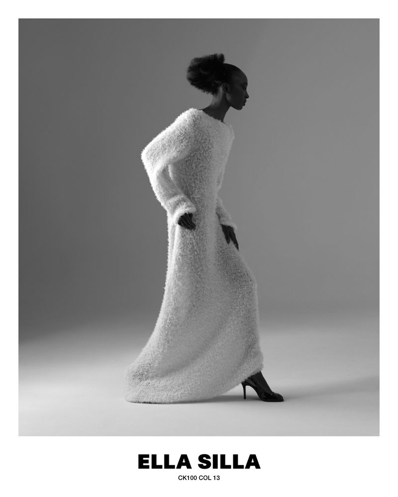 Model wearing a long textured white dress and heels against a gradient grey studio background in a black and white fashion portrait.