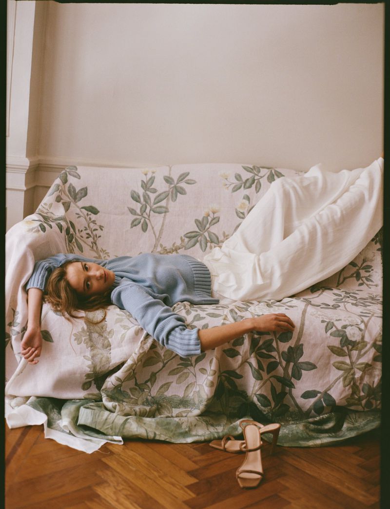 Woman in a light blue sweater and white trousers lying on a patterned floral sofa with strappy heeled sandals on the wooden floor.