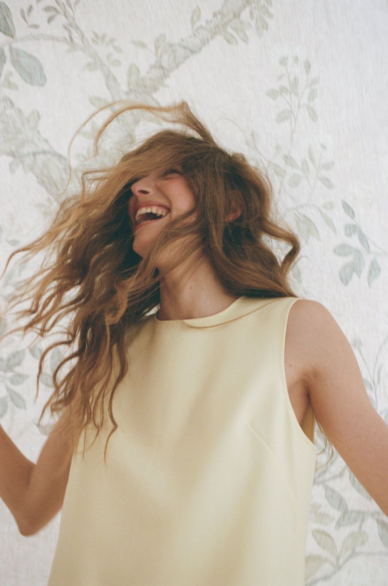 Smiling woman with long blonde hair tossing her head wearing a yellow sleeveless top against a patterned botanical wallpaper.