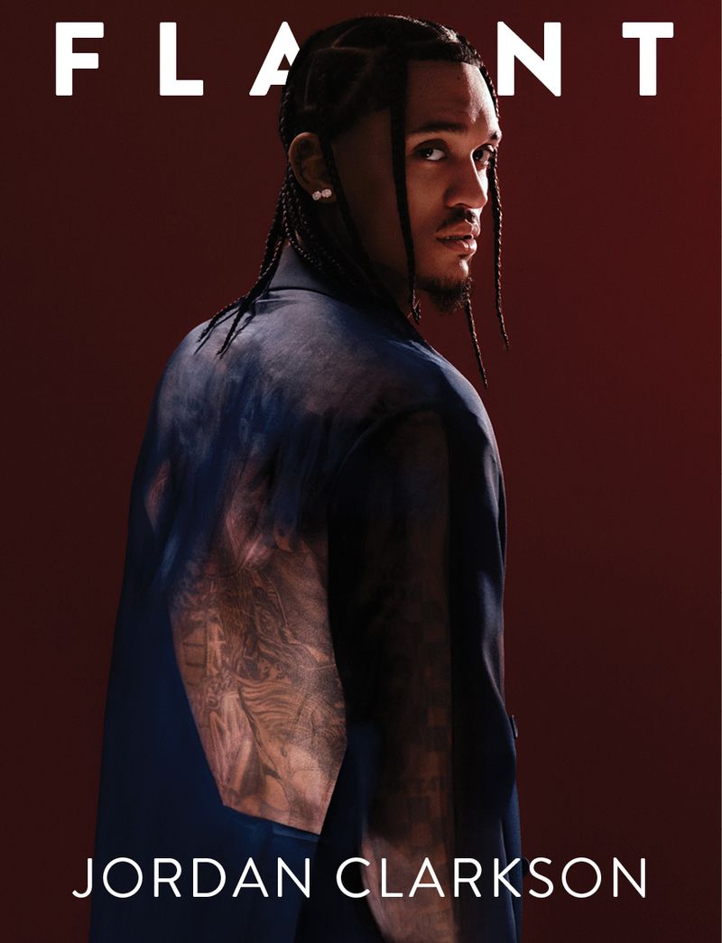 Portrait of Jordan Clarkson on magazine cover, wearing navy blazer, looking over shoulder, with braided hair against dark red backdrop.